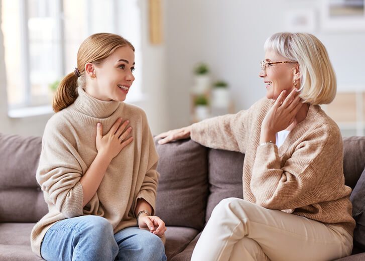 Young woman smiling and talking with senior mother while sitting on comfortable sofa at home together