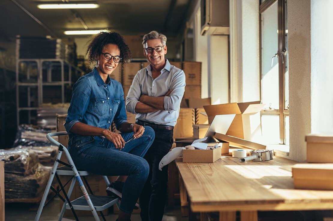 Smiling business partners together at online seller warehouse. Young woman and mature man looking at camera and smiling at drop shipping business office.