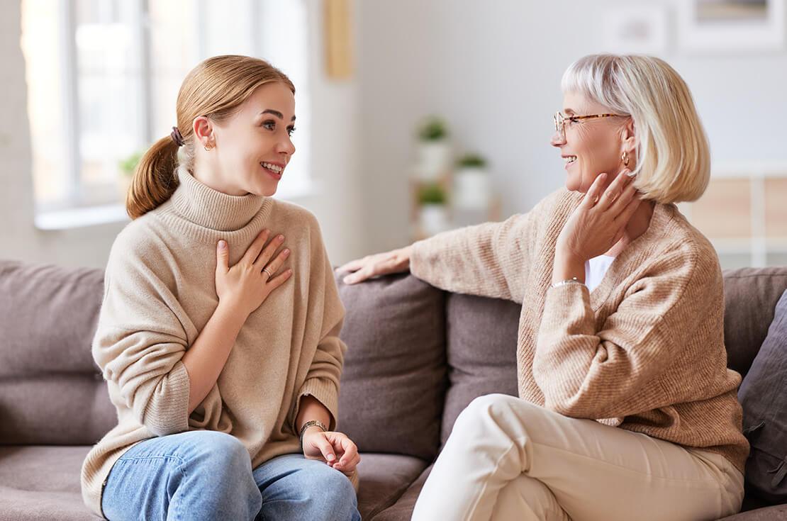 Young woman smiling and talking with senior mother while sitting on comfortable sofa at home together